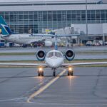 A private jet on the runway at a bustling airport with commercial planes and terminal in view.